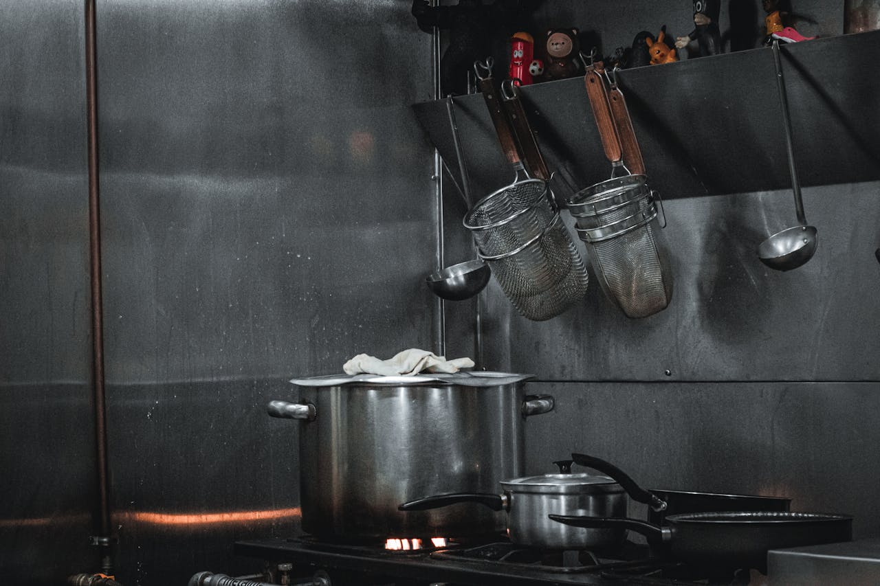 services-01 Stainless steel commercial kitchen setup with pots, strainers, and utensils hanging on a rail.